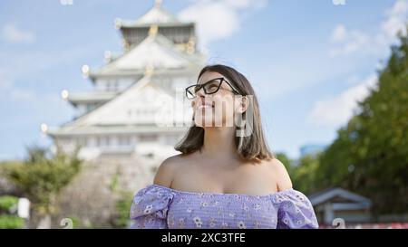 Confident and joyful hispanic woman in glasses, smiling while posing happily at famous osaka castle, her carefree expression reflecting the beauty of Stock Photo
