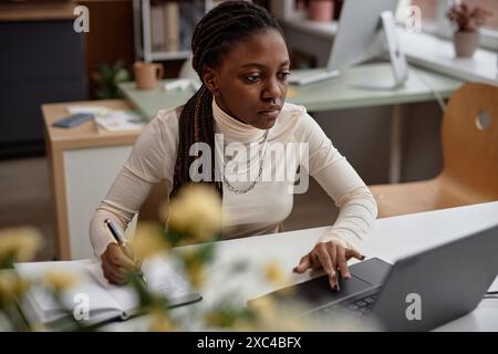 Medium shot of focused young African American woman working on laptop in coworking space, office or classroom. Student preparing for exams, taking notes, self-study concept, copy space Stock Photo
