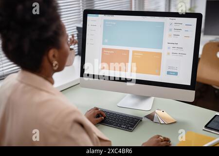 Over shoulder shot of African American woman using computer in office. PC screen displaying online education platform with UI, UX design lessons, focus on monitor, copy space Stock Photo