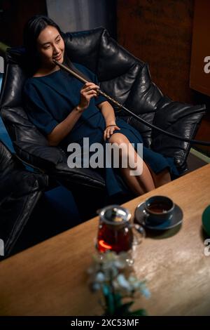 Elegant Asian lady in dress smoking hookah sitting in comfortable armchair Stock Photo