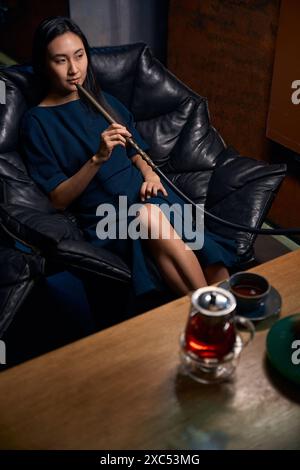 Relaxed Asian woman sitting in leather armchair smoking hookah Stock Photo