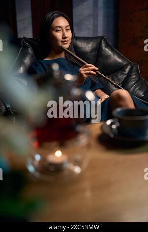 Self-confident attractive woman smoking hookah sitting in big cozy armchair Stock Photo