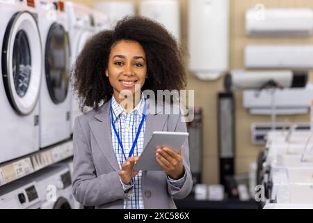 Portrait of a smiling young African American female assistant and sales agent in a business suit standing in a store near a household appliance, holding a tablet and looking at the camera. Stock Photo