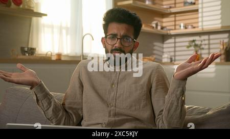 Indian Arabian muslim man in kitchen freelancer businessman working laptop from home puzzled shrugging shoulders don't know male customer confused Stock Photo