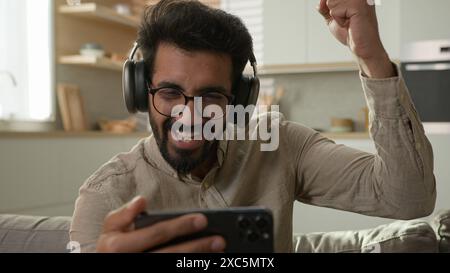 Surprised happy Arabian Indian man in headphones watch video stream on mobile phone excited male home owner watching football match online winning Stock Photo