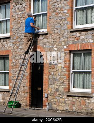 June 2024 - Mature man up a ladder alone cleaning windows beside a road on his own without anyone holding the base of it. Stock Photo