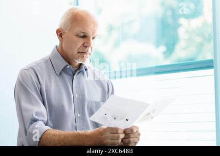 bald man with a white goatee, wearing a light blue shirt, sits by a window reviewing documents. His serious expression suggests he is considering both Stock Photo