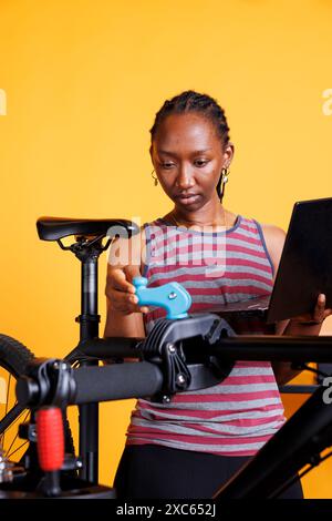 Determined black woman cyclist inspecting, adjusting and repairing damaged bicycle using laptop and specialized tools. Young lady researching for bike maintenance instructions on wireless computer. Stock Photo
