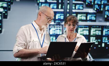Focused IT inspectors looking around data center, using laptop and tablet to crosscheck disaster recovery plan and assess high tech establishment units in need of replacement, preventing perils Stock Photo