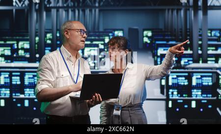 Proficient repairmen looking around high tech data center, using laptop to crosscheck disaster recovery plan and assess server rows in need of replacement, preventing hindrances Stock Photo