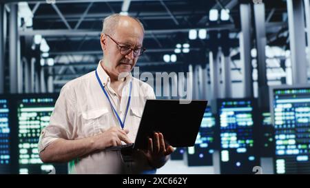 Proficient IT professional navigating network of servers in industrial establishment. Programmer with laptop in hand ensuring seamless operations, doing optimizations and maintenance in data center Stock Photo
