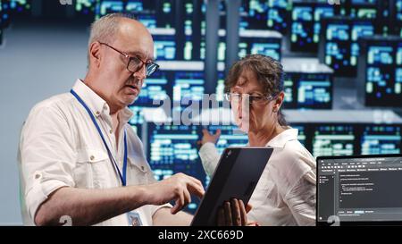 Organized admins in high tech workspace, completing tasks on tablet while running scripts on laptop terminal. Focused specialists amidst server clusters, accessing databases and manipulating code Stock Photo
