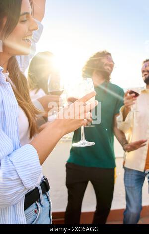 Side view of young Caucasian woman holding wine with lens flare and friends dancing on rooftop. Stock Photo