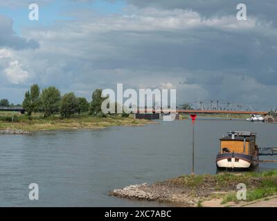 A calm river with a small boat on the bank, grassy trees and a bridge in the background under a cloudy sky, zutphen, gelderland, netherlands Stock Photo