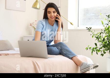 Female freelancer with laptop talking by mobile phone in bedroom Stock Photo