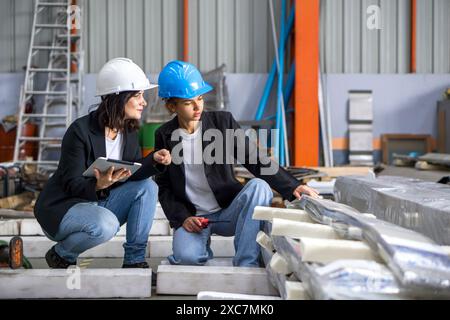 Two workers wearing safety helmet are discussing products that need to be shipped in a factory. One holds a tablet while the other points. Stock Photo