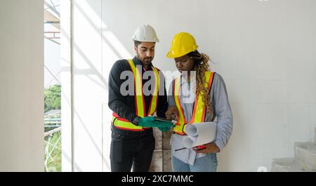 Two construction workers checking work schedule on tablet computer. Wear hardhat and safety vest, stand between stairway in construction site. Stock Photo