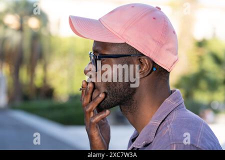 A young African American man smoking a cigarette, wearing a pink cap and glasses, standing outdoors - Casual smoker in profile - Relaxed and candid at Stock Photo