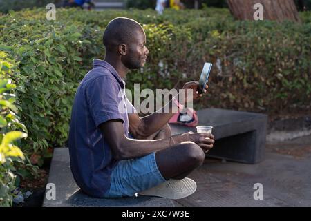 A young African American man sitting outdoors in a park, using a smartphone and holding a drink - Casual and relaxed scene - Modern technology, leisur Stock Photo