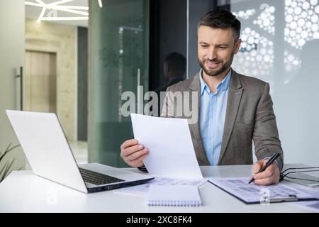 Smiling businessman working at desk with laptop, reviewing documents and taking notes in a modern office Stock Photo