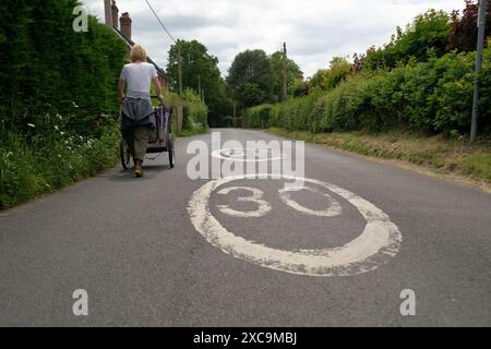 A middle aged woman walks along a country lane in hampshire England  on the left hand side pushing a dog carrier next to a 30mph road sign Stock Photo