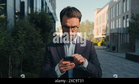 Serious Caucasian entrepreneur adult man businessman CEO business employer office manager outdoors looking at smartphone using mobile phone gadget Stock Photo