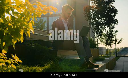 Caucasian business man freelancer work with laptop computer male businessman office worker on steps modern university campus in sunny city browsing Stock Photo
