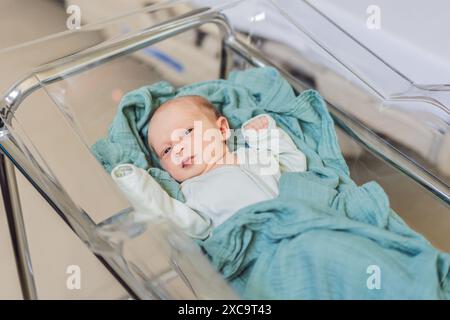 A newborn rests peacefully in his transparent bassinet in the hospital. The clear bassinet provides visibility for medical staff to monitor the baby's Stock Photo