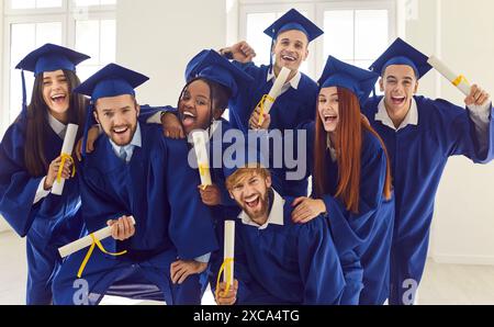 Group of laughing happy multiracial graduates hugging indoors holding diploma. Stock Photo