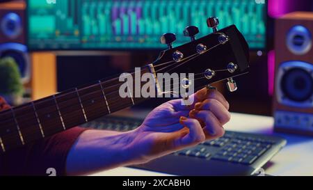 Musical performer tuning his guitar by twisting the knobs, preparing to play acoustic instrument in home studio. Artist recording sounds to create new track, using equalizer mixing gear. Camera A. Stock Photo