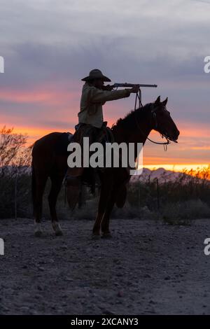 Sunset silhouette of a cowboy pointing a rifle, sitting on his horse. Stock Photo