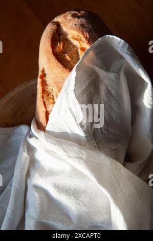 A loaf of fresh french bread wrapped in a white linen tea towel in a wooding bowl on a wooden farm table. Stock Photo