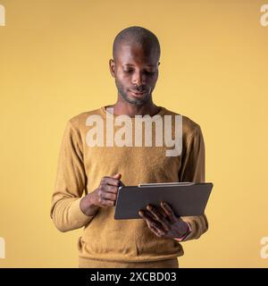 A young African American man using a tablet, standing casually on a yellow background - Casual tech use - Modern and focused atmosphere, digital lifes Stock Photo