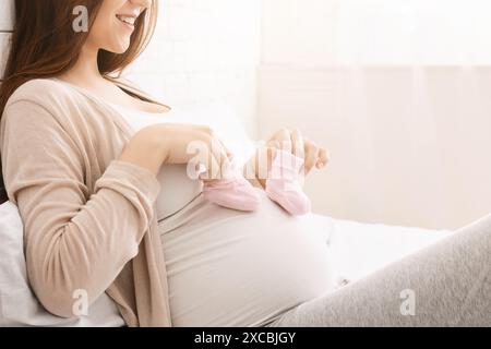 Pregnant Woman Holding Baby Shoes While Smiling in Bright Room Stock Photo