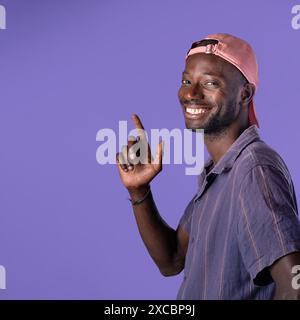 A cheerful young African American man wearing a pink cap and casual shirt, smiling and pointing up on a purple background - Happy and positive portrai Stock Photo