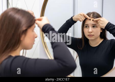 Young, beautiful woman with long hair looks in the mirror, squeezing a pimple on her forehead. Stock Photo