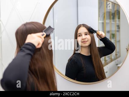 Young woman in a black long sleeve brushes her long hair with a hairbrush while looking in the mirror. Stock Photo