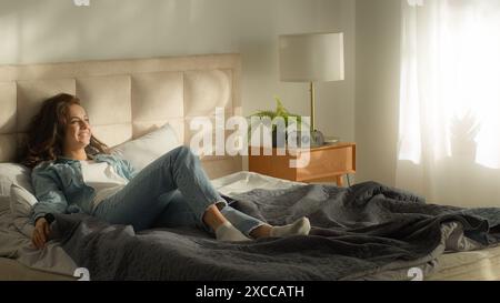 Happy couple woman lying resting man jumping on bed homeowners home love boyfriend girlfriend together relationship laughing enjoyment fun cozy Stock Photo