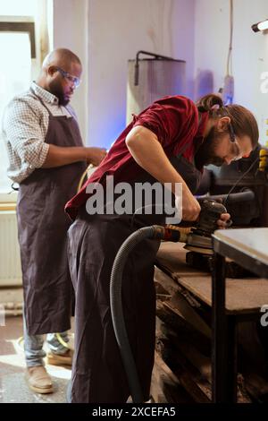 Carpenter and african american coworker using angle grinder on lumber block to smooth surfaces. Man uses orbital sander machinery for furniture assembling, helped by apprentice in background Stock Photo