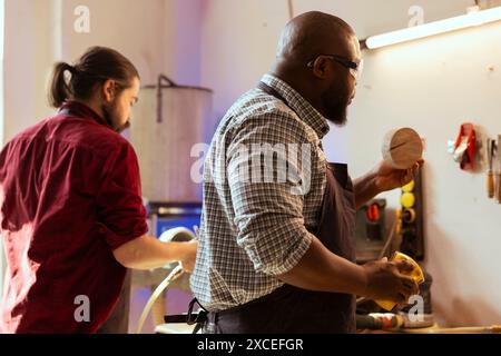 African american manufacturer doing last touches on piece of wood before using it for furniture assembly, sanding with sandpaper. BIPOC woodworking professional examining timber block for damages Stock Photo