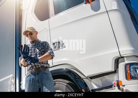 A truck driver, wearing a plaid shirt and a baseball cap, is putting on his blue work gloves in front of his white semi-truck. Stock Photo