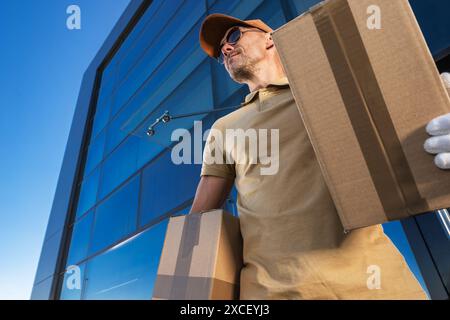 Delivery guy carrying two large cardboard boxes, he looks up, possibly searching for an address. This image represents the efficiency and reliability Stock Photo