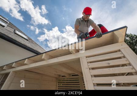 A man wearing a red hat and safety glasses is working on the roof of a wooden structure. He is using a power drill to secure a piece of roofing materi Stock Photo