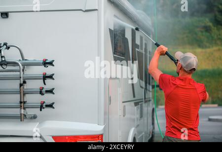 A man in a red shirt washes the side of a white motorhome with a pressure washer. The motorhome has a bike rack on the back and is parked in an outdoo Stock Photo