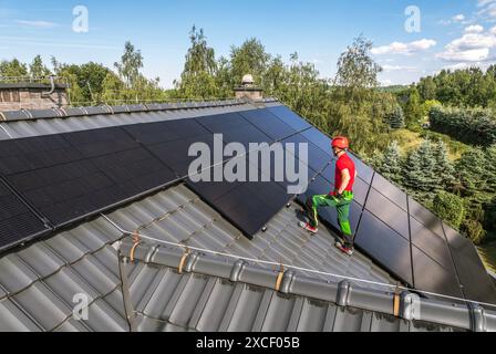 A worker in a red hard hat and green pants is installing solar panels on a roof. The panels are black and are laid out in a grid pattern. The roof is Stock Photo