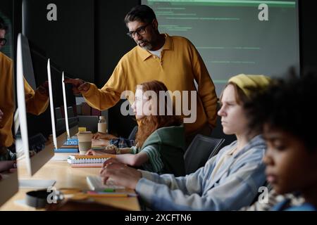 Mature biracial Computer Science teacher helping Caucasian girl to find mistake in code during lesson in modern classroom Stock Photo