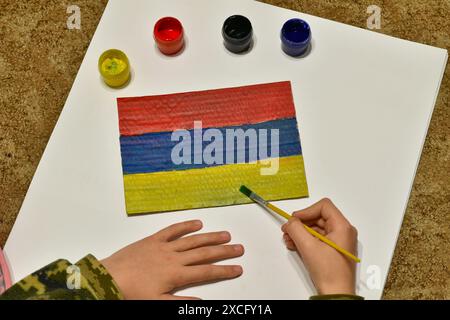 The boy on the floor draws the flag of Armenia with a brush and paints on paper. Stock Photo