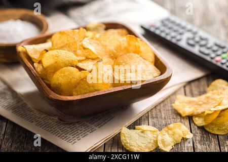 A bowl filled with potato chips sits on a wooden table, alongside a television remote . The focus is on the bowl of chips, which are piled high and lo Stock Photo