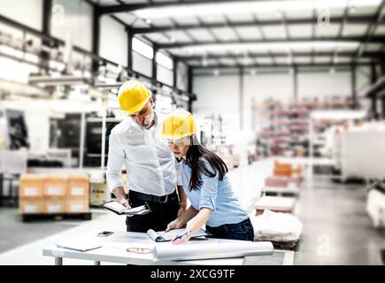 Two project managers standing in modern industrial factory, looking blueprints and documents on table. Manufacturing facility with robotics, robotic Stock Photo