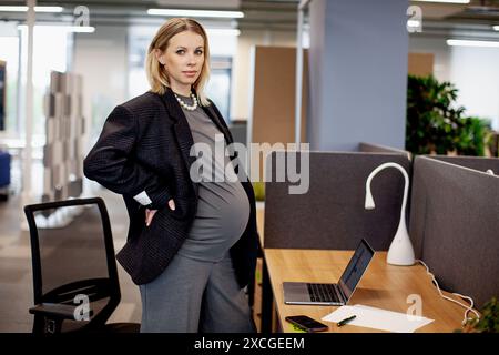 Pregnant woman standing in office cubicle, surrounded by a desk, computer, chair, and office supplies. Portrait of professional pregnant businesswoman Stock Photo